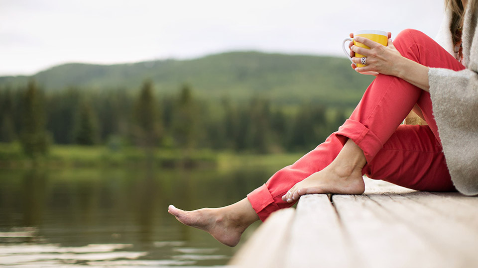 A barefoot woman sitting on a dock by a calm lake holding a mug.